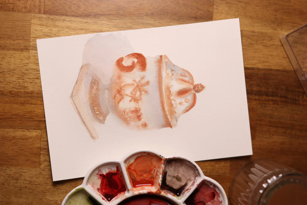 Photograph from above of a watercolor of a lidded jar in terracotta tones on paper, with a palette of red, orange and brown paint and a glass of water on a wooden table.