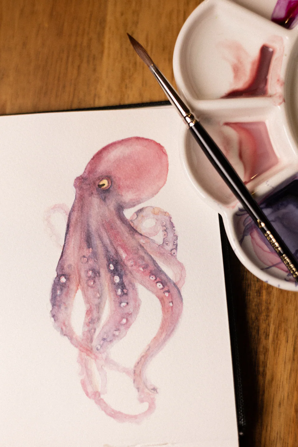 Photograph from above of a watercolor octopus on a sheet of paper, with a round brush and a white palette holding rose and violet paint, on a wooden table.